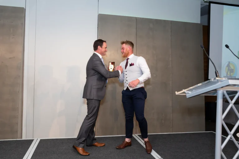 This photograph captures a moment of recognition at the Pearses 50th Anniversary Awards Night at Zinc, Fed Square. On stage, the award recipient, wearing a white shirt, gray patterned vest, burgundy tie, and navy trousers, shakes hands with the presenter in a gray suit and striped tie, holding a small framed award in his left hand. Both men smile warmly at each other. The stage features a dark gray carpet, a clear podium with microphones, and modern gray and white paneled walls, creating a formal yet celebratory atmosphere of congratulations and achievement.