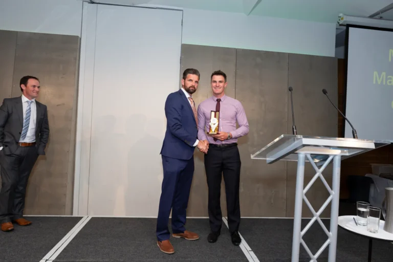 This photograph captures a formal moment at the Pearses 50th Anniversary Awards Night at Zinc, Fed Square, as an award recipient shakes hands with the presenter on stage. The recipient, wearing a light purple shirt, burgundy tie, and black pants, holds a small silver trophy and smiles at the camera. The presenter in a blue suit also smiles. A third man in a gray suit and striped tie stands to the side, observing. The stage features a modern gray and white paneled backdrop, a clear podium with microphones, and a small table with a pitcher and glasses, highlighting the celebratory awards atmosphere.