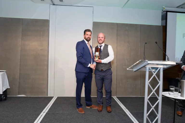 This photograph, taken at the Pearses 50th Anniversary Awards Night at Zinc, Fed Square, captures a celebratory moment as a man in a white shirt and gray plaid vest receives a trophy from a presenter in a blue suit. Both are smiling and shaking hands on stage. The background features a gray paneled wall, a clear podium, and a small table with glasses of water, highlighting the formal and festive atmosphere of the awards presentation.