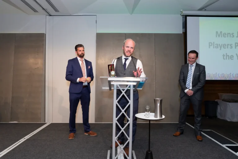 This photograph captures a celebratory moment at the Pearses 50th Anniversary Awards Night at Zinc, Fed Square, as a bearded man in a gray vest and white shirt delivers a speech at a clear podium, gesturing enthusiastically. A small award is visible on the podium. He is flanked by two men in suits, smiling and looking on with admiration. The stage features a gray paneled backdrop and a projection screen displaying part of the award title, "Mens J Players Pl the Yea." The scene conveys excitement, recognition, and camaraderie during the awards presentation.
