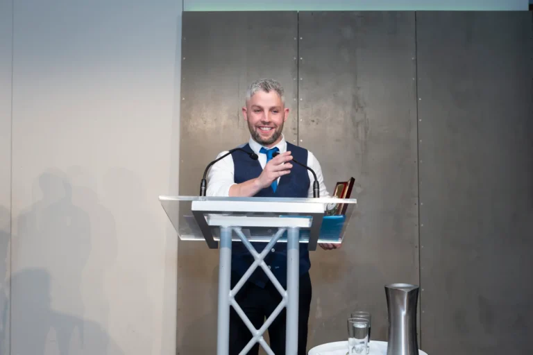This photograph, taken during the Pearses 50th Anniversary Awards Night at Zinc, Fed Square, captures a man delivering an enthusiastic awards speech on stage. The speaker, with a short beard and graying hair, is dressed in a white shirt, navy vest, and blue tie. He gestures with his right hand while holding a small award in his left hand on a clear acrylic podium with two microphones. A small table with a metal jug and glass is nearby. The modern gray-paneled backdrop and bright stage lighting emphasize the celebratory and formal atmosphere of the event.