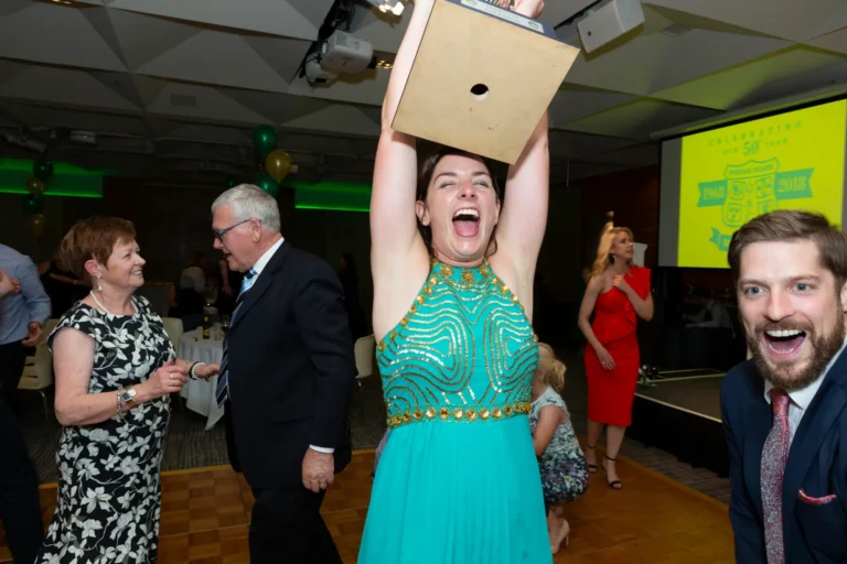 The image captures a thrilling moment of triumph at the Pearses 50th Anniversary Awards Night. In the center, a woman in a shimmering teal halter-neck dress raises a large square trophy high above her head, her face lit with pure joy and excitement. Beside her, a man in a dark suit and patterned tie reacts dramatically toward the camera with an open-mouthed expression of surprise and delight, adding humor and energy to the scene. Behind them, smiling guests and clusters of green and gold balloons create a festive backdrop. The wooden dance floor and projected anniversary logo complete the lively, celebratory setting.
