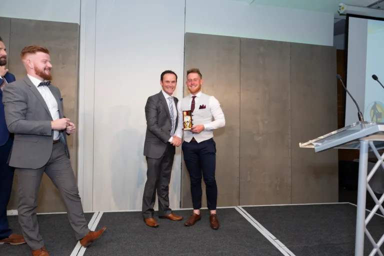 This photograph captures a celebratory moment at the Pearses 50th Anniversary Awards Night at Zinc, Fed Square. On stage, the award recipient, dressed in a white shirt, gray vest, and navy trousers, holds a small gold-colored trophy while shaking hands with the presenter in a dark gray suit. Both smile at the camera, while a third man in a gray suit and bow tie approaches from the side. The stage features dark gray carpeting, a clear podium with a microphone, and gray paneled walls, creating a formal and joyful atmosphere during the awards presentation.