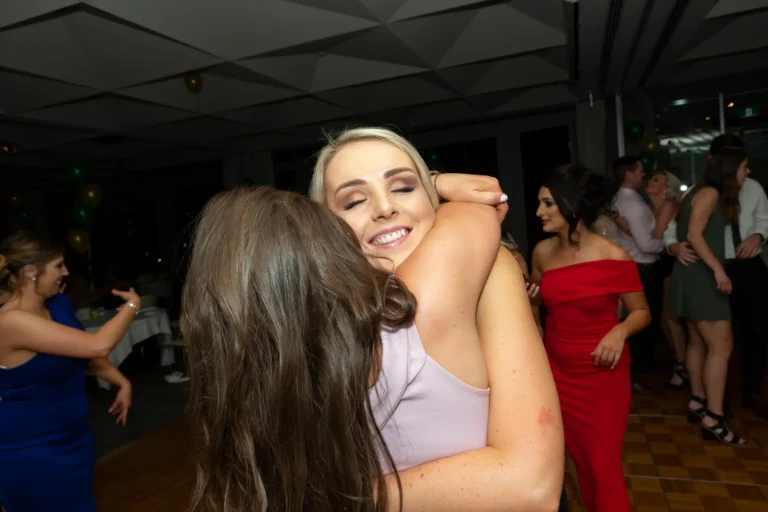 A heartfelt moment at the Pearses 50th Anniversary Awards Night shows two women embracing on a wooden dance floor. The woman in a light pink dress smiles with her eyes closed as she hugs a friend in a dark dress. In the background, guests in red and blue dresses dance and mingle, while clusters of green and gold balloons decorate the modern event space. The atmosphere is joyful and festive, capturing friendship, affection, and candid celebration amidst the lively awards night.
