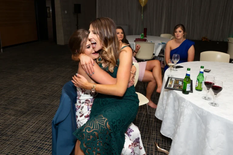 A joyful and candid moment at the Pearses 50th Anniversary Awards Night. A woman in a dark green lace dress sits on the lap of a friend in a white floral dress, hugging and laughing while her friend smiles supportively. They are seated at a table with wine glasses, beer bottles, and a framed photo. Other guests are visible at nearby tables, and a cluster of balloons hangs above. The setting is a formal event space with a dark patterned carpet, capturing a lively and social celebration.