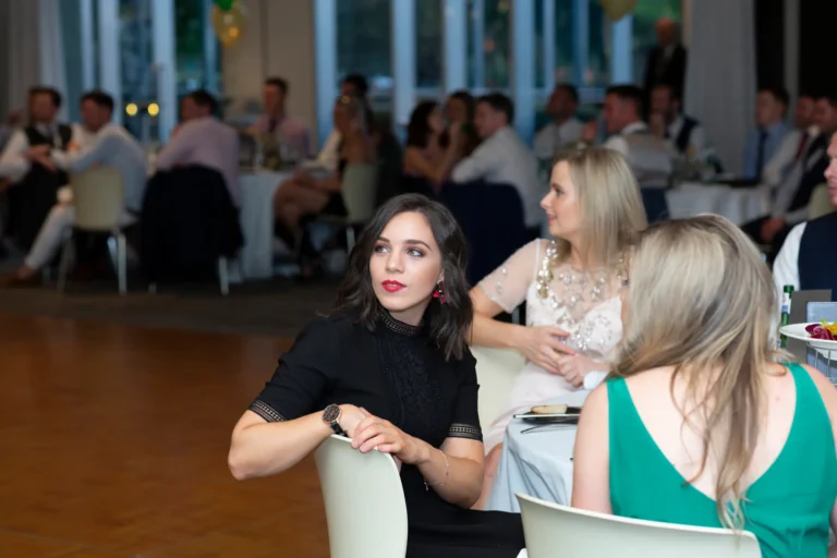 A candid moment at the Pearses 50th Anniversary Awards Night. A woman with dark hair and red lipstick, wearing a black lace high-neck dress, sits in a white chair looking thoughtfully to the left. Beside her, a blonde woman in a light embellished dress and another guest in a green dress are seated. The table has glasses and cutlery. The background shows a formal event space with round tables, guests in suits and dresses, gold and green balloon clusters, and a wooden floor with large windows. The atmosphere is social and relaxed.