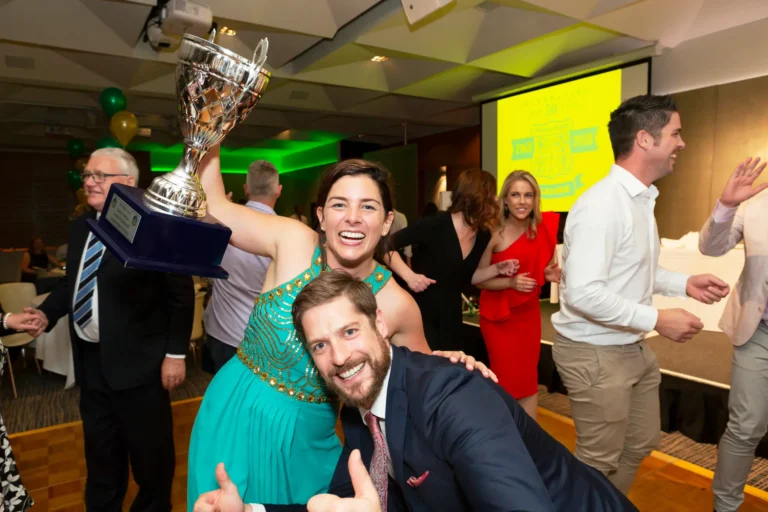 The image captures a jubilant and energetic scene at the Pearses 50th Anniversary Awards Night. In the foreground, a bearded man in a dark suit smiles broadly and gives a thumbs-up to the camera. Behind him, a woman in a glittering teal dress proudly raises a large silver trophy above her head, her face glowing with triumph and excitement. Around them, guests in formal attire cheer and smile — a man claps enthusiastically, another raises his hands in celebration, and a woman in a red dress beams in the background. The dance floor and anniversary logo projection complete the lively, festive atmosphere.