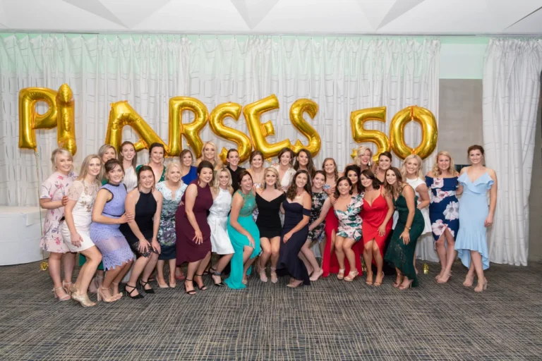 The image captures a large group of women posing together at the Pearses 50th Anniversary Awards Night. Arranged in several rows, some women stand while others kneel in front, all smiling at the camera. They wear a variety of formal dresses in different colors, styles, and patterns. Behind them, a large gold balloon sign reads "PEARSES 50," emphasizing the milestone celebration. The indoor venue features a pleated light-colored curtain backdrop and a patterned carpet. The atmosphere is festive, joyful, and social, capturing a memorable group moment.