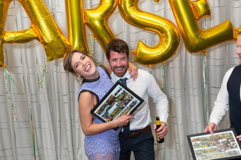The image captures a joyful and candid moment at the Pearses 50th Anniversary Awards Night. A woman in a light purple lace-patterned dress laughs warmly as she leans toward a bearded man beside her, who is smiling broadly and has his arm around her. The man, wearing a white dotted shirt with a dark tie, holds a bottle of beer while both look fondly at a framed photo collage in the woman’s hands. In the background, large gold balloons hang against a light gray curtain, adding to the festive and celebratory atmosphere of the evening.