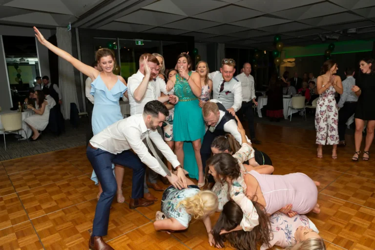 A lively group of men and women celebrate on the dance floor at the Pearses 50th Anniversary Awards Night. Some are standing, including a woman in a ruffled light blue dress with her arm raised triumphantly, while others are playfully piled or crouched on the floor, smiling and laughing. Men in white shirts and women in colorful dresses, including teal and floral patterns, share joyful expressions. The wooden dance floor, tables with guests in the distance, and clusters of green and gold balloons create a festive and energetic atmosphere, capturing a candid, fun, and celebratory moment during the event.