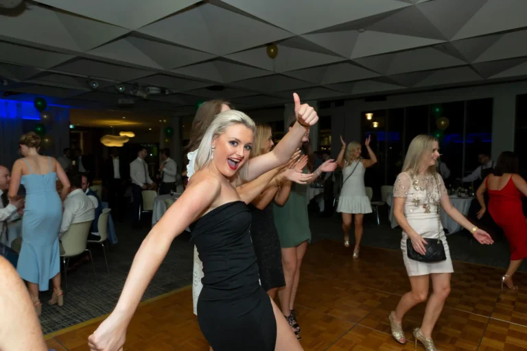 The image captures a joyful and energetic scene from the Pearses 50th Anniversary Awards Night. In the foreground, a woman with blonde hair in a black strapless dress beams with excitement, extending her arms and giving a thumbs-up as she dances. Behind her, several women join in the fun — one in a dark green dress clapping along, and another in a sparkling light-colored dress dancing joyfully. The wooden dance floor is surrounded by round tables draped in white tablecloths, where guests are mingling and watching. Gold and green balloons decorate the elegant venue, enhancing the festive, post-awards celebration atmosphere.