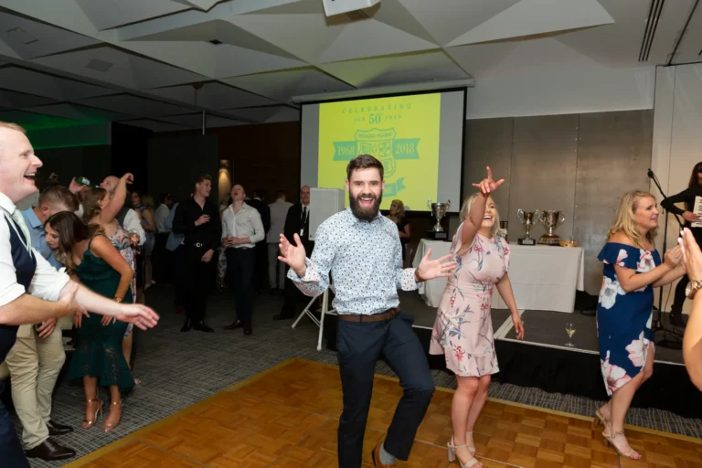 A lively and energetic moment on the dance floor at the Pearses 50th Anniversary Awards Night. A bearded man in a polka-dot shirt dances with arms outstretched, smiling widely, while a woman in a light pink floral dress dances beside him with one arm raised. Other guests in the background are also dancing and smiling. A stage with a projection screen displays "CELEBRATING OUR 50 YEARS 1968-2018," with trophies lined up on a table. The wooden dance floor and festive crowd create a joyful, celebratory atmosphere, capturing the excitement and happiness of the anniversary event.