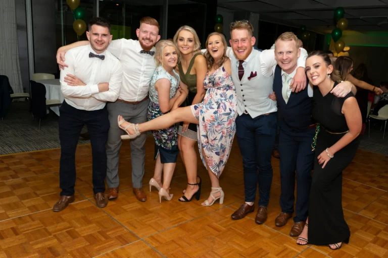 A lively group of eight people—four men and four women—pose together on the dance floor at the Pearses 50th Anniversary Awards Night. Arms around each other, they smile and look at the camera, with one woman playfully raising her leg. Men wear formal suits and ties, while women wear a mix of patterned and solid dresses. The festive event space features a dark wood dance floor, clusters of green and gold balloons, and round tables with white tablecloths. The candid moment captures camaraderie, celebration, and joyful energy among friends at the awards night.