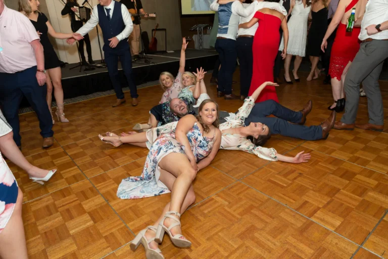 A lively, playful moment on the wooden dance floor at the Pearses 50th Anniversary Awards Night. A group of people, mainly women, are lying and sitting in a fun pile-up, smiling and laughing. A woman in a floral dress lies on her side in the foreground, while a man with a beard raises his arms in celebration. Other women around them are smiling and raising their arms. In the background, guests are standing and dancing, and a live band with an accordion and guitar performs on a stage with a screen showing the event logo.