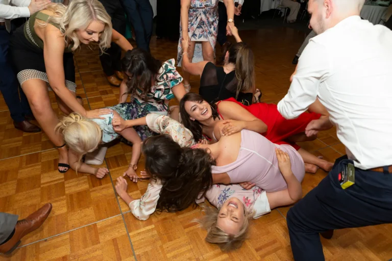 A lively and joyful group of people, mostly women, are playfully piled on a wooden dance floor at the Pearses 50th Anniversary Awards Night. Women in light purple, red, floral, and dark green dresses laugh energetically, while a man in a white shirt and blue pants leans over, smiling at the group. Everyone is engaged in a spontaneous, celebratory moment, radiating happiness and fun. The background shows a spacious event room with a wooden floor, highlighting the group’s playful interaction and capturing the vibrant, festive atmosphere of the 50th anniversary celebration.