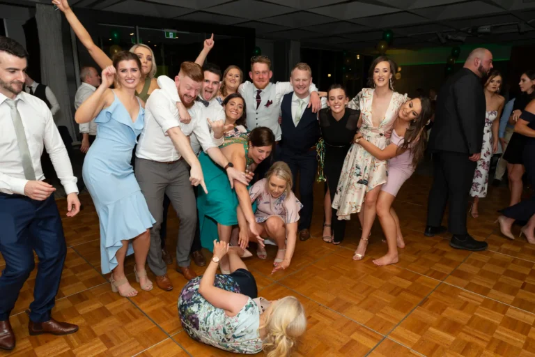 A large group of men and women pose energetically on the dance floor at the Pearses 50th Anniversary Awards Night. In the foreground, a woman in a floral dress lies laughing, while others bend, squat, and lean toward the camera, smiling and pointing. Men in suits and women in formal dresses share playful expressions. The wooden checkered dance floor and clusters of green and gold balloons set a festive backdrop, capturing the joyful, lively, and celebratory atmosphere as the group enjoys a playful moment together during the party portion of the event.
