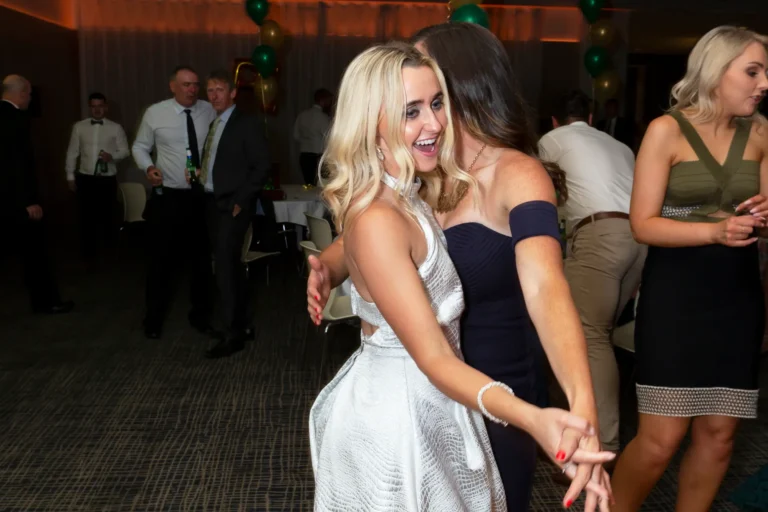 A joyful moment on the dance floor at the Pearses 50th Anniversary Awards Night shows two women dancing together. The blonde woman in a light-patterned halter dress laughs with her mouth open, holding hands with her friend in a dark blue strapless dress. Guests mingle and dance in the background, including men in white shirts and ties. The room features a dark patterned carpet, white tables and chairs, and clusters of green and gold balloons. The bright lighting enhances the lively, celebratory atmosphere, capturing the energy and happiness of the 50th anniversary celebration.