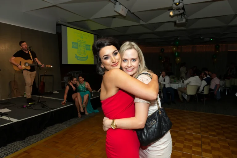 A lively, celebratory moment at the Pearses 50th Anniversary Awards Night shows two women embracing on a wooden dance floor. The woman in a strapless red dress smiles confidently at the camera, while her friend in a light-colored lace dress hugs her warmly, holding a quilted black purse. In the background, a band performs on stage, a large screen displays the event logo "CELEBRATING OUR 50 YEAR 1968–2018," and guests at round tables watch. Green and gold balloons hang from the ceiling, creating a joyful, festive atmosphere during the awards night.