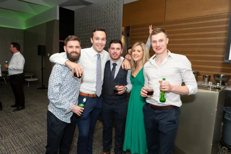 A group of five people, three men and two women, pose for a lively group photo at the Pearses 50th Anniversary Awards Night. They stand close together with arms around each other, smiling warmly at the camera. The men are dressed in button-down shirts, with one wearing a suit vest and tie, while the women wear a green dress and a patterned outfit. Several hold drinks, including a green beer bottle. The background shows a bar area, a speaker setup, and other guests mingling in a modern, textured wooden room with patterned carpeting, creating a festive and social atmosphere.