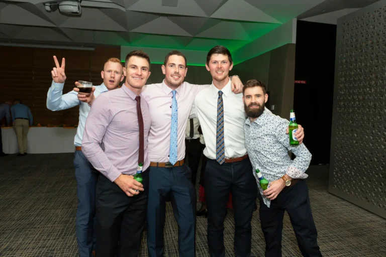 A group of five men posing for a celebratory group photo at the Pearses 50th Anniversary Awards Night. They are standing close together with their arms around each other’s shoulders, smiling at the camera. The men are dressed in formal attire, holding drinks, and appear joyful. The background shows a formal event space with a dark patterned carpet and a table in the distance, creating a festive and social atmosphere.
