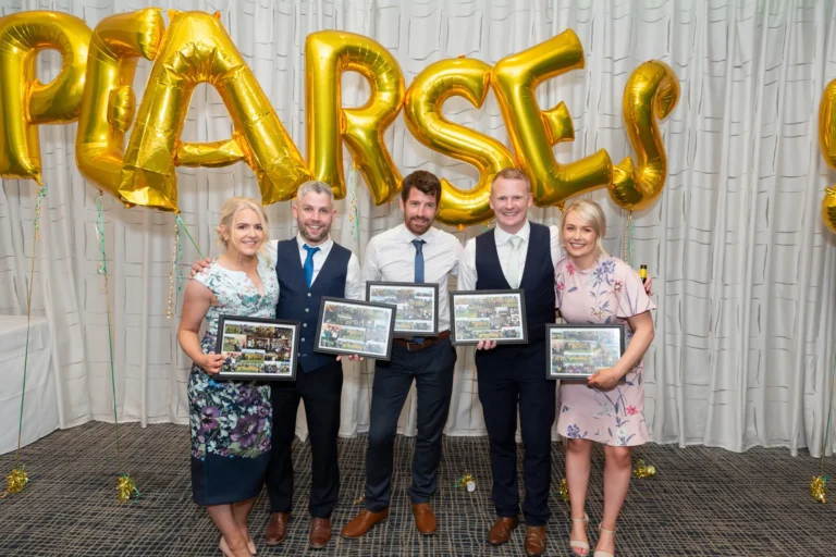 A group of five people—three men and two women—pose for a celebratory photo at the Pearses 50th Anniversary Awards Night. They stand in a line with arms around each other, smiling at the camera. Several hold framed awards or photos. The background features a white pleated curtain with large gold balloons spelling "PEARSES." The dark patterned carpet is scattered with gold confetti, and a table with a white tablecloth is partially visible, highlighting the festive, joyful, and commemorative atmosphere of friendship and celebration at the event.