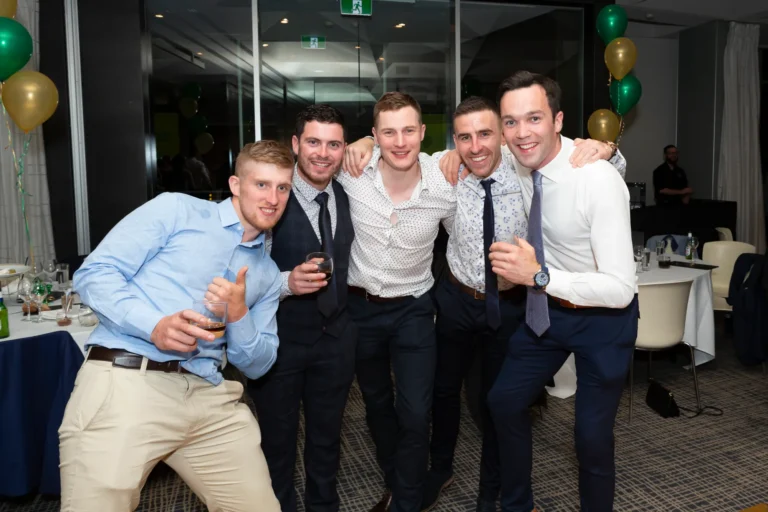 A group of five men poses for a fun and celebratory photo at the Pearses 50th Anniversary Awards Night. They stand close together with arms around each other’s shoulders, smiling broadly at the camera. The man on the far left crouches playfully, holding a drink and giving a thumbs-up, while the others wear suits and ties, some holding drinks. The background shows a formal event space with round tables, white tablecloths, chairs, and clusters of green and gold balloons. The dark patterned carpet and large windows add to the festive and joyful atmosphere of camaraderie and celebration.
