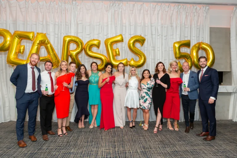 A large group of twelve men and women poses for a celebratory group photo at the Pearses 50th Anniversary Awards Night. The men are dressed in dark suits with ties, and the women wear formal dresses in red, dark blue, teal, light blue, and white. Several people hold drinks, including wine glasses and bottles. They stand close together, smiling at the camera, conveying friendship and camaraderie. The background features a pleated white curtain with gold foil balloons spelling "PEARSES 50," and gold streamers scattered across the dark patterned floor, enhancing the festive and joyful atmosphere.