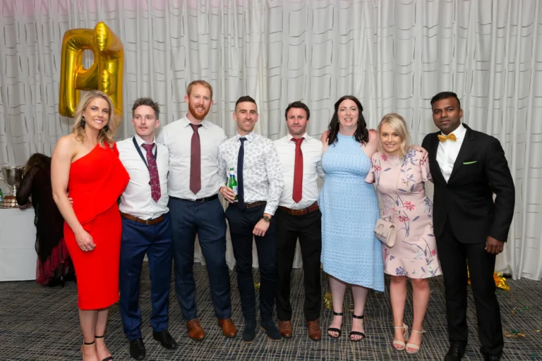 A group of seven people, four men and three women, poses for a festive photo at the Pearses 50th Anniversary Awards Night. They stand in a line, smiling confidently at the camera. The men wear suits, ties, and one a gold bow tie, while the women are in formal dresses in red, light blue, and pink floral. One man holds a green beer bottle. The background shows a white pleated curtain with a large gold foil "P" balloon, a table with trophies, and gold and green streamers scattered on the dark patterned carpet, capturing a joyful moment of camaraderie and celebration.
