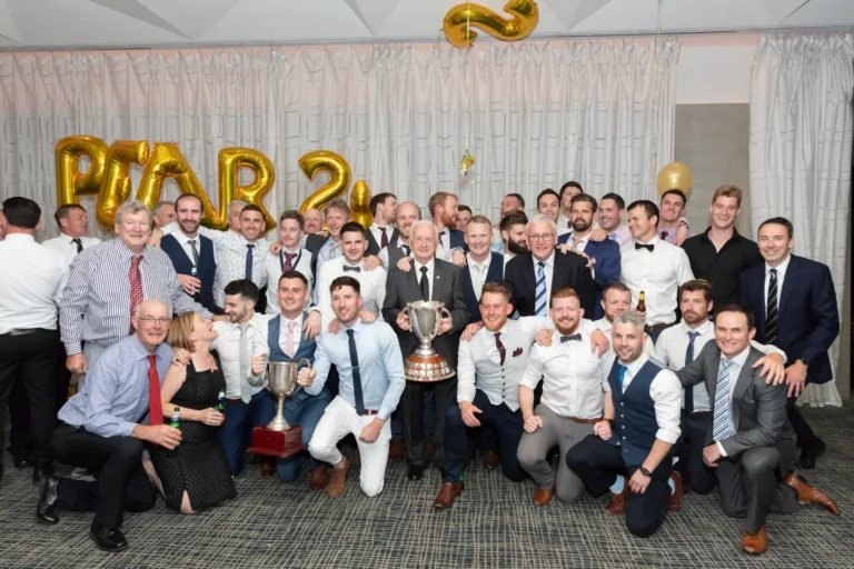 The image captures a large group of men and a few women posing for a celebratory group photo at the Pearses 50th Anniversary Awards Night. Arranged in several rows, some stand while others kneel or crouch in front. Most are smiling and looking at the camera. In the center, two men hold large silver trophies. The men wear suits, vests, or button-down shirts with ties or bow ties, and the women wear formal black dresses. A pleated light-colored curtain and gold balloons in the background highlight the festive, milestone celebration.