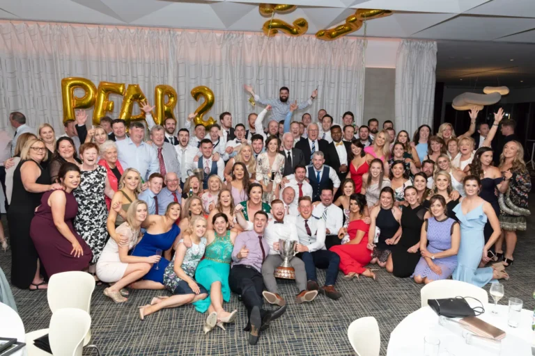 A large, diverse group of men and women pose for a celebratory photograph at the Pearses 50th Anniversary Awards Night. Arranged in several rows, some are standing while others kneel or crouch in front, all smiling at the camera. Two men in the center hold a large silver trophy. Guests wear a mix of formal and semi-formal attire, including suits, vests, button-down shirts, and various dresses. Behind them, a light-colored pleated curtain and a gold balloon sign spelling "PEARSE 50" highlight the festive milestone celebration. The floor is patterned, and round tables are visible in the foreground.