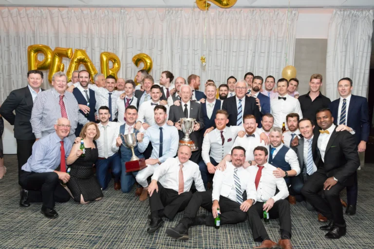 A large group of men and a few women pose for a celebratory photograph at the Pearses 50th Anniversary Awards Night. Arranged in several rows, some are standing while others kneel or crouch in front, all smiling at the camera. Two men in the front hold large silver trophies. The men wear suits, vests, or button-down shirts with ties or bow ties, while the women wear formal black dresses. A pleated light-colored curtain and a gold balloon are in the background, highlighting the festive milestone celebration. The floor is patterned, adding to the formal event setting.