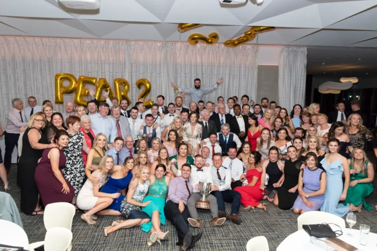 A large, diverse group of men and women pose for a celebratory photograph at the Pearses 50th Anniversary Awards Night. Some are standing while others kneel or crouch in front, all smiling at the camera. Two men in the center hold a large silver trophy. Guests wear formal and semi-formal attire, including suits, vests, button-down shirts, and dresses of various colors and styles. Behind them, a light-colored pleated curtain and a gold balloon sign spelling "PEARSES 50" highlight the milestone celebration. The floor is patterned, with round tables visible in the foreground.