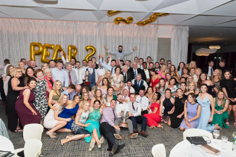 A large, diverse group of men and women pose for a celebratory photograph at the Pearses 50th Anniversary Awards Night. Some are standing while others kneel or crouch in front, all smiling at the camera. Two men in the center hold a large silver trophy. Guests wear formal and semi-formal attire, including suits, vests, button-down shirts, and dresses in various colors and styles. Behind them, a light-colored pleated curtain and a gold balloon sign is visible, along with additional gold balloons. The floor is patterned, with round tables visible in the foreground.