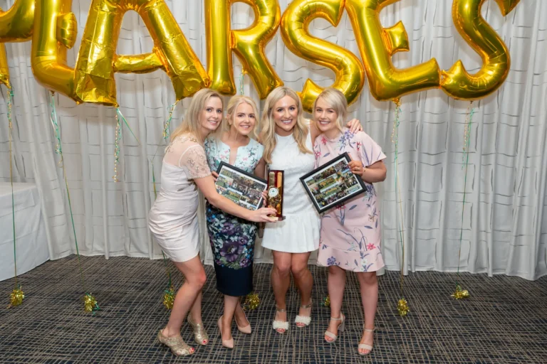 Four women pose for a celebratory group photo at the Pearses 50th Anniversary Awards Night. Two women in the center hold framed awards or mementos while all are smiling at the camera. They are dressed in elegant formal attire, including floral and light-colored dresses, and wearing high heels. The background features a white curtain with large gold balloons spelling "PEARSES," and the dark carpeted floor is scattered with gold confetti, highlighting the festive and joyful atmosphere of camaraderie and celebration at the event.
