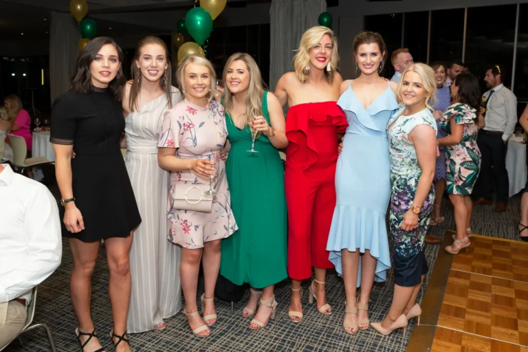A group of seven women poses for a celebratory photo at the Pearses 50th Anniversary Awards Night. They stand in a line, smiling confidently at the camera, dressed in a variety of formal outfits including dresses, a jumpsuit, and floral patterns. One woman in a green dress holds a drink. The background shows other guests socializing and seated at tables, with clusters of gold and green balloons adding to the festive atmosphere. The dark patterned carpet and wooden dance floor contribute to the lively setting, capturing a joyful moment of friendship and celebration.