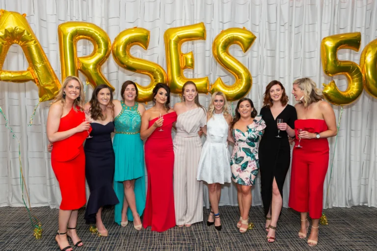 A group of eight women poses together at the Pearses 50th Anniversary Awards Night, smiling warmly at the camera and holding wine glasses. They are dressed in a mix of formal attire, including red, navy, teal, floral, and black dresses, as well as a light gray jumpsuit. The pleated white curtain backdrop features large gold foil balloons spelling part of "PEARSES 50," with gold confetti scattered on the dark patterned floor. The image captures a joyful, festive moment of friendship, celebration, and camaraderie during the 50th-anniversary event.