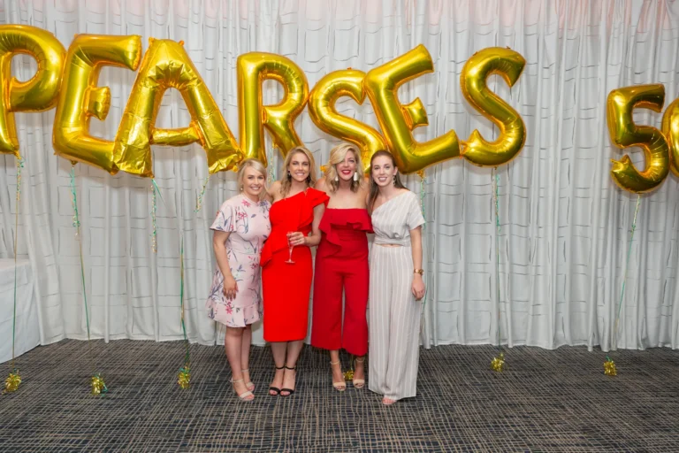 A group of four women poses together at the Pearses 50th Anniversary Awards Night, smiling warmly at the camera. They are dressed in formal attire: a light pink floral dress, two red dresses (one holding a wine glass), and a cream-colored top with wide-leg pants. The backdrop features a pleated white curtain and large gold foil balloons spelling "PEARSES 50." Gold confetti and streamers decorate the dark patterned floor. The image captures a joyful, festive moment of friendship and celebration, highlighting camaraderie and the elegant, celebratory atmosphere of the event.