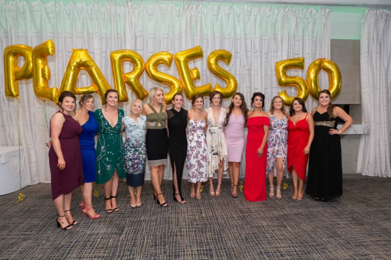A group of women stand side by side, smiling and posing for a celebratory group photo at the Pearses 50th Anniversary Awards Night. They wear a mix of colorful formal dresses and jumpsuits, some with floral or patterned designs. Behind them, large gold balloons spell "PEARSES 50," with gold confetti scattered on the dark carpeted floor. Their joyful expressions and confident poses convey friendship, camaraderie, and the festive spirit of the milestone celebration, capturing a lively and memorable moment of shared happiness at the event.