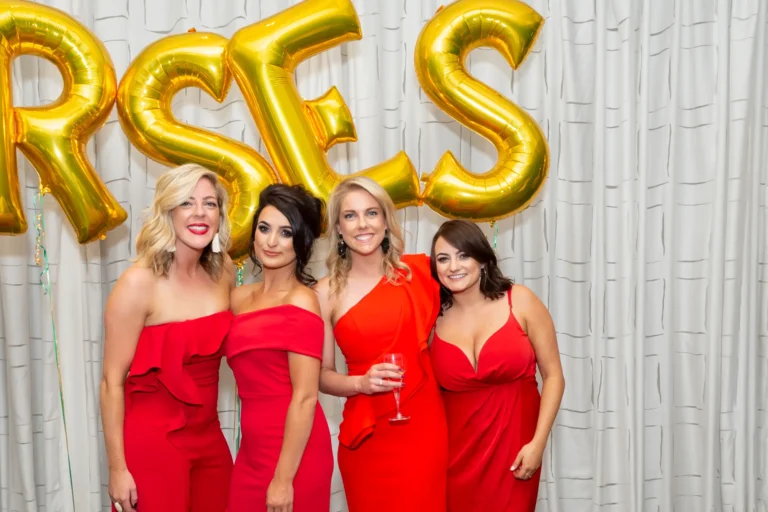 A group of four women poses together for a celebratory photo at the Pearses 50th Anniversary Awards Night. They are all wearing red dresses in different styles, smiling warmly and looking at the camera. One woman holds a wine glass. Behind them, a white pleated curtain and large gold foil balloons spelling "RSES" form a festive backdrop. The coordinated outfits, bright smiles, and decorative elements create a joyful, elegant, and celebratory atmosphere, highlighting the camaraderie and festive spirit of the event.