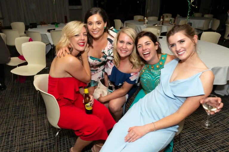 The image captures a lively and celebratory moment of five women posing together at the Pearses 50th Anniversary Awards Night. Two women are seated on chairs while the other three kneel or stand behind them. They are all smiling at the camera, dressed in colorful formal attire including red, floral, blue, green, and light blue dresses. Several hold drinks, including a bottle and a glass of white wine. The background shows a large event room with round tables set with white tablecloths, glasses, and cutlery. The atmosphere is joyful and social.