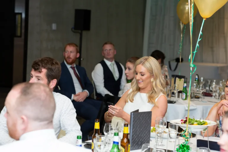 A candid moment at the Pearses 50th Anniversary Awards Night. A woman with long blonde hair in a white sleeveless dress sits at a round table, smiling faintly while looking at her phone. Other guests, including men in a blue suit, white shirt, and dark vest, are seated around her, engaged with the event. The table holds beer bottles, glasses, and dinner plates. The background shows a formal dining hall with white tablecloths, green and gold balloon clusters, gray paneled walls, and a large window. The atmosphere is festive yet attentive.