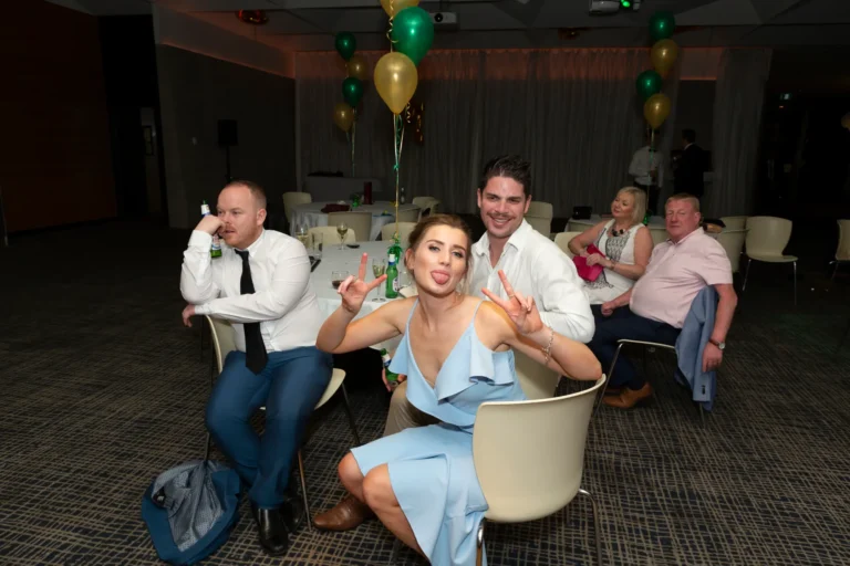 A playful, candid moment at the Pearses 50th Anniversary Awards Night shows a woman in a light blue ruffled dress seated in a chair, sticking out her tongue and flashing peace signs at the camera. A bearded man behind her smiles with his arm around her. Other guests at the table, including men in white shirts and a blonde woman in white, are engaged and smiling. The formal event space features round tables with glasses and bottles, white chairs, dark patterned carpet, and green and gold balloons hanging from the ceiling, creating a festive and lively atmosphere.