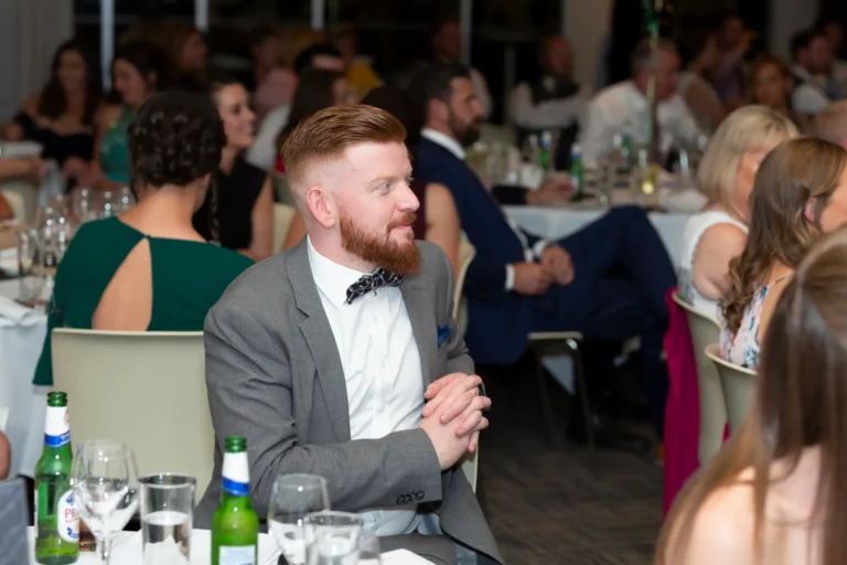 A candid moment at the Pearses 50th Anniversary Awards Night. A man with a reddish beard and modern haircut, wearing a gray suit, white shirt, and dark patterned bow tie, sits at a round table with hands clasped, looking thoughtfully to his right. The table has wine glasses, beer bottles, and water glasses. Other formally dressed guests are visible in the background, including a woman in a green dress and a man in a blue suit. The warm lighting and dark patterned carpet create a relaxed, social, and elegant atmosphere.