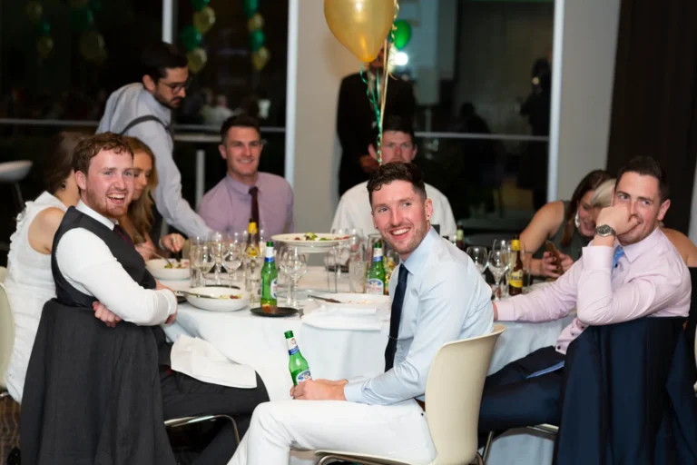 A lively, candid moment at the Pearses 50th Anniversary Awards Night. A man in a light blue shirt and dark tie smiles widely at the camera, holding a green beer bottle, while two men beside him, one in a white shirt and vest and another in a pink shirt, laugh and enjoy the moment. Guests are seated around a table set with plates, glasses, and bottles. The background shows a formal event space with large windows, a high ceiling, and green and gold balloon clusters, creating a festive and joyful atmosphere.
