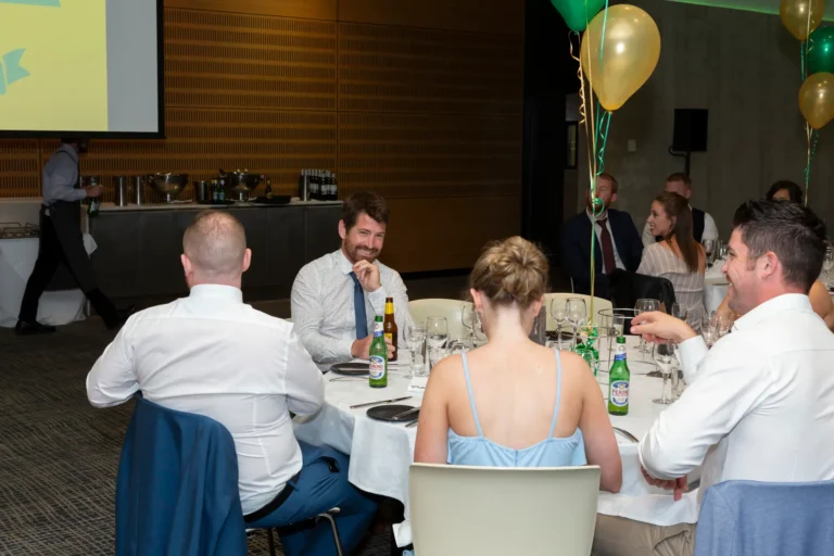 The image captures a candid and cheerful moment at the Pearses 50th Anniversary Awards Night, showing guests seated around a round table engaged in conversation. A man with a beard, wearing a white shirt and blue tie, sits at the center, smiling and looking to his left, with a beer bottle in front of him. Across from him, a woman in a light blue dress faces away from the camera. Two other men in white shirts are visible, one gesturing animatedly. The background features another table of guests, a man walking past, and festive gold and green balloons adding a lively, celebratory atmosphere.