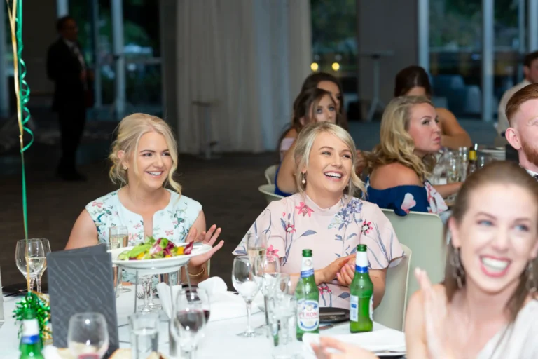 Guests react with joy during the Pearses 50th Anniversary Awards Night at Zinc, Fed Square. In the foreground, a group of women seated at a round table with white tablecloths are laughing and smiling, engaging with each other. The table holds wine glasses, bottles, and plates of food. In the background, other guests watch the event, seated at tables decorated with green balloons and streamers. The high-ceilinged venue with large windows creates an elegant, lively atmosphere, capturing the warmth and celebratory spirit of the awards ceremony.