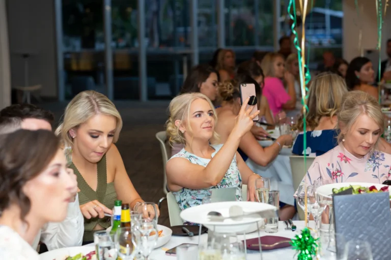 A candid moment at the Pearses 50th Anniversary Awards Night at Zinc, Fed Square, shows a blonde woman in a floral dress holding up her phone to capture a photo or video of the event. She sits at a round table set with dinner plates, wine glasses, and bottles. Other guests at the table are engaged in their own activities. The background features a modern event space with large windows, a high ceiling, and green and gold balloons and streamers, highlighting the lively and celebratory atmosphere of the awards night.
