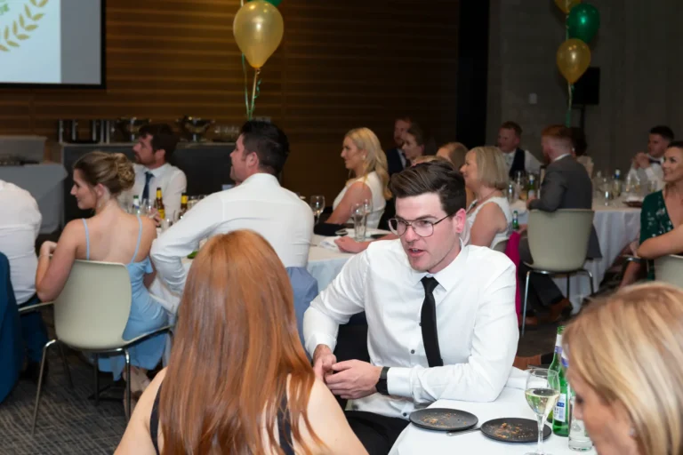 The image captures a candid and engaging moment at the Pearses 50th Anniversary Awards Night. In the foreground, a man with glasses, wearing a white shirt and black tie, leans forward as he speaks animatedly to a woman with long red hair, who is seated with her back to the camera. His expression is lively, suggesting a friendly and engaging conversation. Behind them, other guests are seated at round tables covered in white tablecloths, chatting or looking toward the front of the room. The elegant event space features gray paneled walls, a projection screen, and festive green and gold balloon decorations hanging from the ceiling.