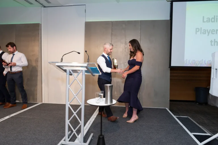 This photograph, taken during the Pearses 50th Anniversary Awards Night at Zinc, Fed Square, captures a formal moment of recognition on stage. A man standing behind a podium, dressed in a suit, smiles as he shakes hands with a woman in a dark blue off-the-shoulder gown walking toward him to receive her award. Another man is visible in the background holding a bottle. The stage features gray-paneled walls, a large screen, and a small side table with a glass of water. Bright lighting highlights the celebratory and professional atmosphere of the awards presentation.