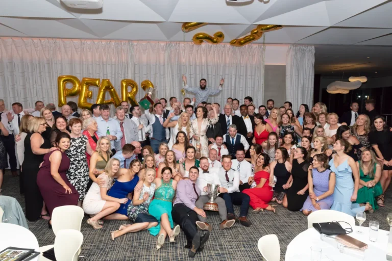 A large, festive group of men and women of various ages pose for a celebratory photograph at the Pearses 50th Anniversary Awards Night. Many are standing while others kneel, crouch, or sit on the floor, all smiling at the camera. Two men in the center hold a large silver trophy. Guests wear formal and semi-formal attire, including suits, vests, shirts, and dresses in various colors and styles. Behind them, a light-colored pleated curtain and a gold balloon sign spelling "PEARSES 50" are visible, along with other gold balloons. The floor is patterned, with round tables partially visible in the foreground.